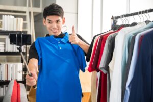 A smiling man holds up a blue polo shirt and gives a thumbs up while taking a selfie, highlighting the importance of customer reviews. He stands beside a rack of assorted clothes in a bright, modern room.