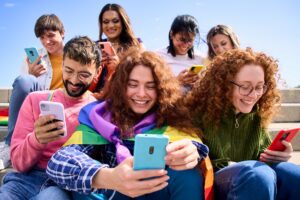 A group of smiling young people sit on outdoor steps, looking at their phones and exploring social media channels. Two in front wear rainbow flags, and everyone appears happy and engaged under a clear blue sky.