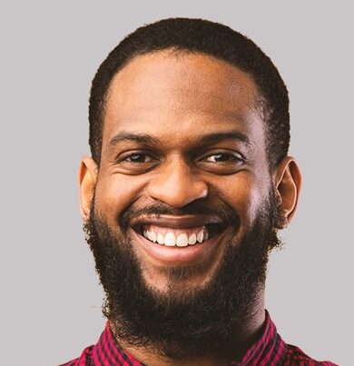 A man with short, dark hair and a beard smiles warmly at the camera. He is wearing a red and black plaid shirt with the collar buttoned, and is posed against a plain, light gray background.