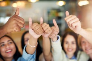 A group of people smiling and giving thumbs up gestures in a bright indoor setting, symbolizing positive customer testimonials. The focus is on their hands, with faces slightly blurred in the background.